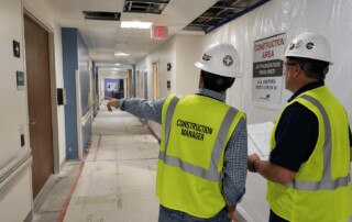 Construction managers reviewing plans inside an active hospital corridor renovation with containment barriers, exposed ceiling panels, and restricted work-zone controls.