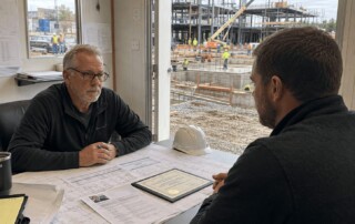 Construction hiring leader reviewing a candidate resume and certification documents inside a jobsite trailer with active building construction visible outside.