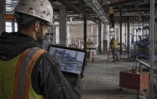 VDC/BIM manager reviews a 3D coordination model on a laptop inside an active commercial construction site with exposed MEP systems.